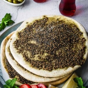 Flatbread covered with zaatar spice and oil on a plate with a side of sliced tomatoes and lemon.