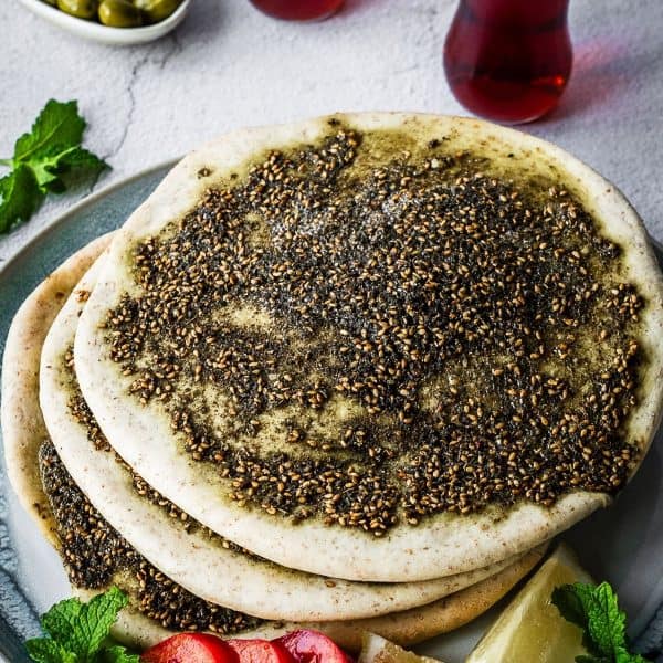 Flatbread covered with zaatar spice and oil on a plate with a side of sliced tomatoes and lemon.