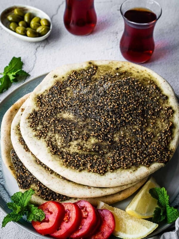 Flatbread covered with zaatar spice and oil on a plate with a side of sliced tomatoes and lemon.