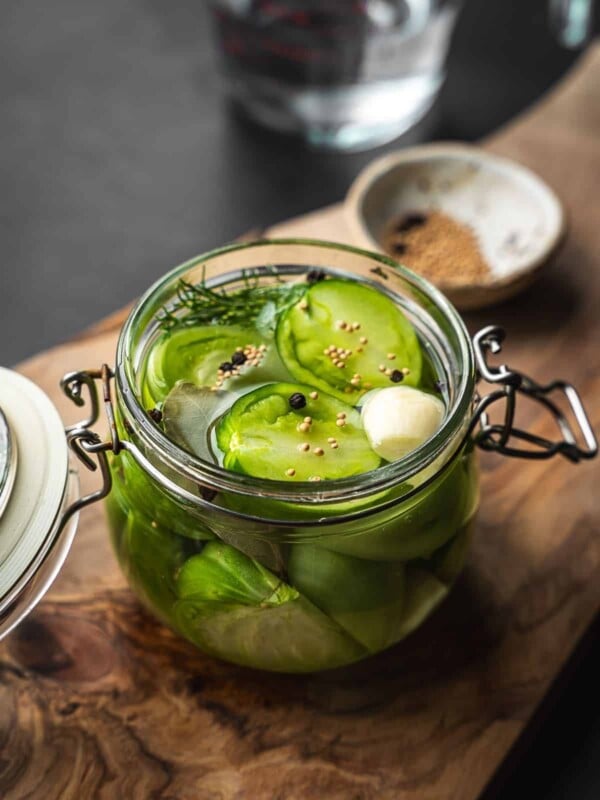 Overhead shot of a jar of pickled sliced green tomatoes and spices on a wooden board.