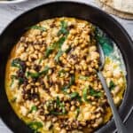 Bowl of warm chickpeas and tahini, garnished with parsley and pine nuts served with sliced cucumbers and pita.