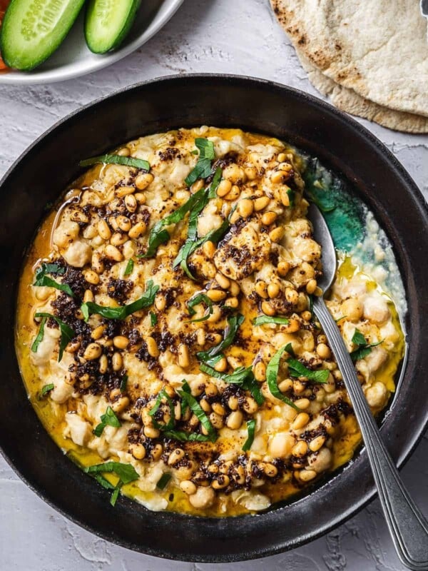 Bowl of warm chickpeas and tahini, garnished with parsley and pine nuts served with sliced cucumbers and pita.