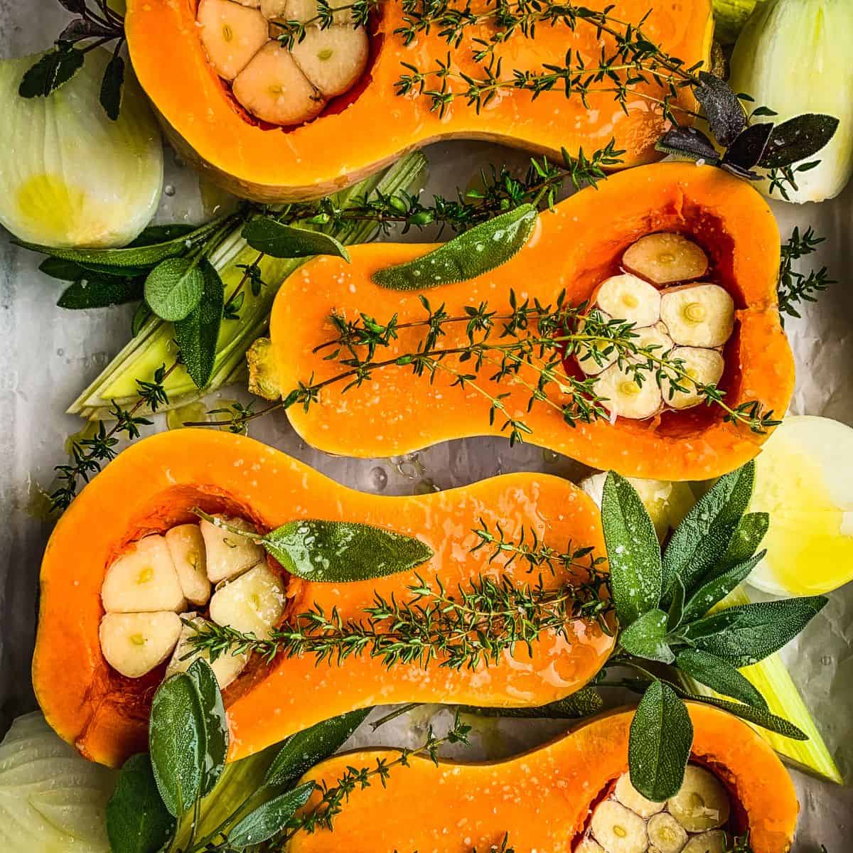 A baking tray with butternut squash cut down the middle and garnished with garlic, leeks and sage.