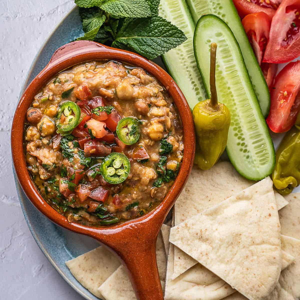 Plate of stewed fava beans with sliced cucumbers, tomatoes and pita bread.