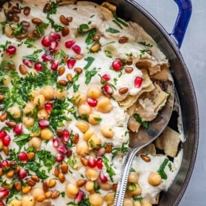 A casserole dish with creamy fattet hummus, garnished with fresh parsley, pine nuts and pomegranate seeds.