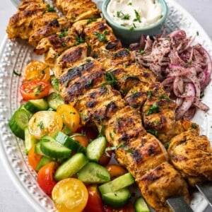 A plate of shish tawook skewers with a side salad, pickled sumac onions and garlic sauce.