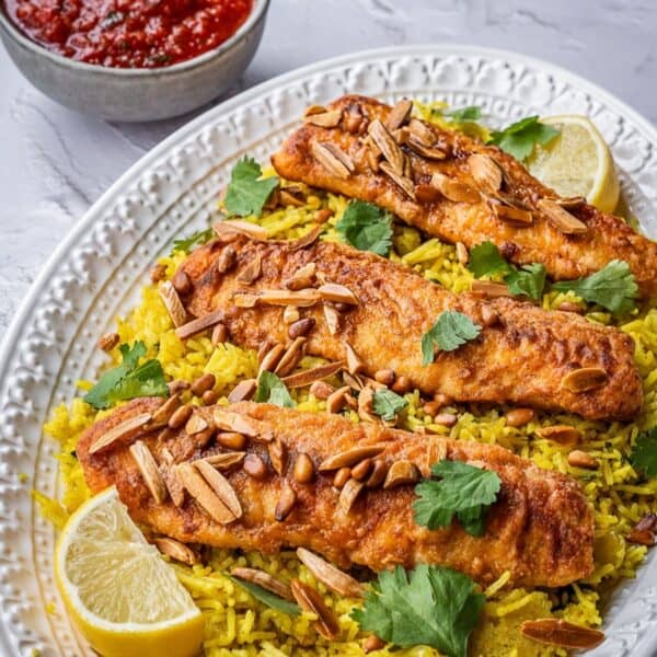Plated fish filets over rice on a serving platter garnished with fried nuts and fresh cilantro with a bowl of tomato sauce in the back.