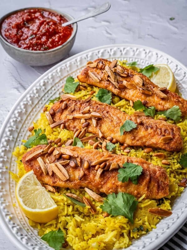 Plated fish filets over rice on a serving platter garnished with fried nuts and fresh cilantro with a bowl of tomato sauce in the back.