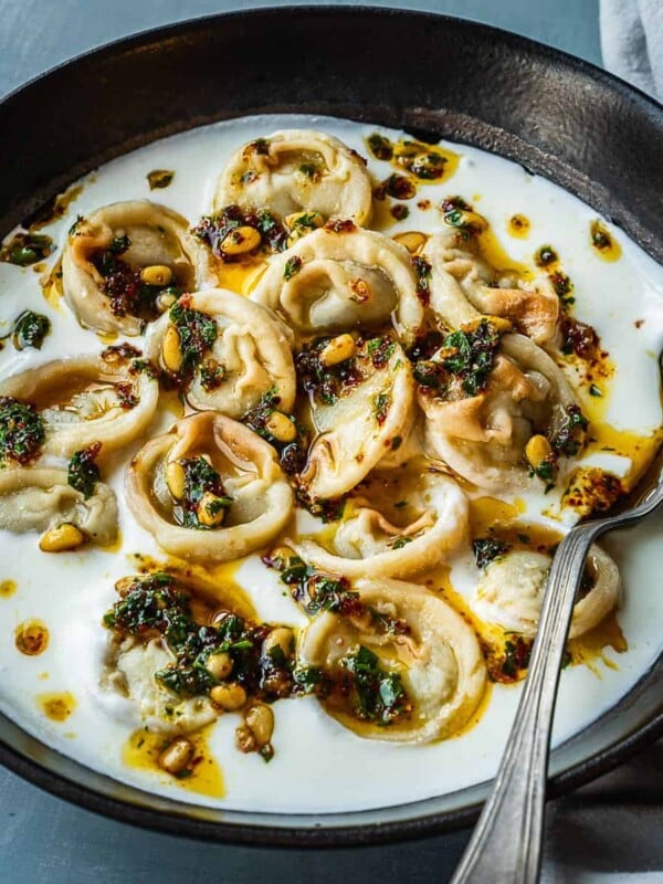 Bowl of shish barak garnished with pine nuts and parsley.