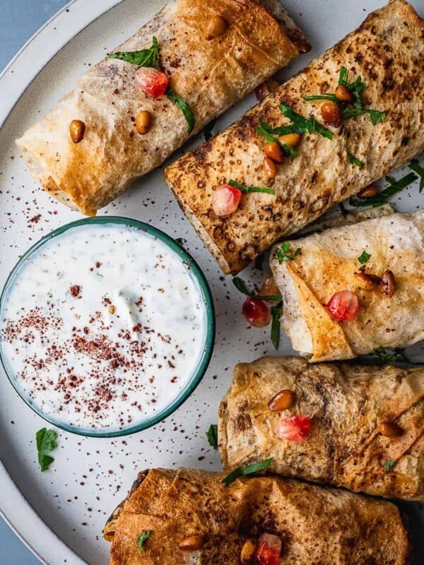 Plate with musakhan rolls with a bowl of mint yogurt, garnished with pomegranate seeds and pine nuts.