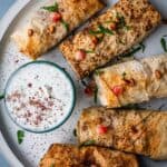 Plate with musakhan sumac chicken rolls with a bowl of mint yogurt, garnished with pomegranate seeds and pine nuts.