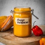 A jar of mango habanero hot sauce on a board with a mango and chile peppers.