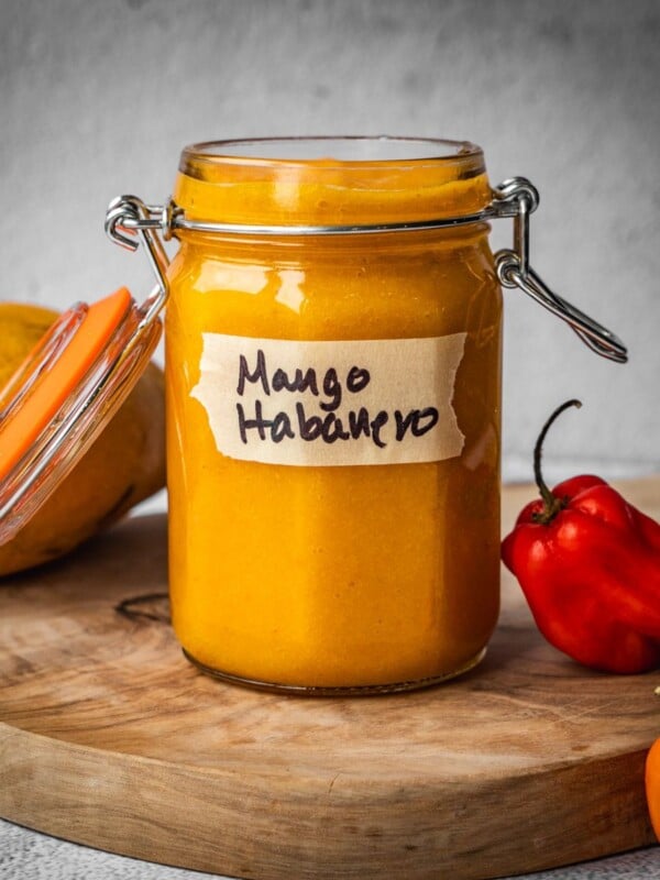 A jar of mango habanero hot sauce on a board with a mango and chile peppers.