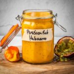 Passion fruit habanero in a jar with a fresh habanero pepper and an opened passion fruit on a board.