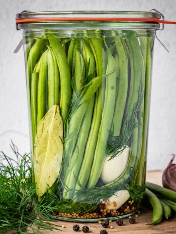 Sealed and labeled jar of dilly beans on a board with dill, peppercorns, mustard seeds, beans and garlic around the base.