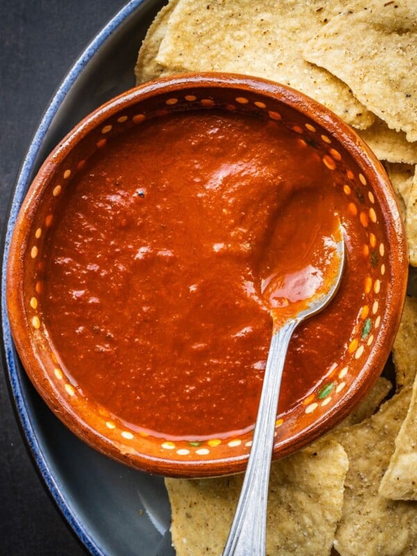 Salsa roja in a terracotta bowl with a spoon on a plate with some tortilla chips.