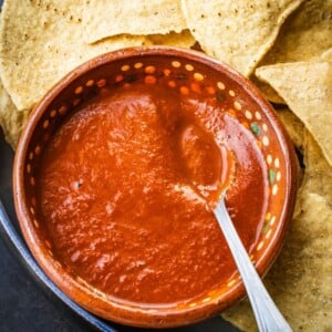 Salsa roja in a terracotta bowl with a spoon on a plate with some tortilla chips.