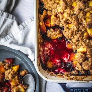 Blueberry peach crisp in a rimmed baking tray with a potion on a plate.