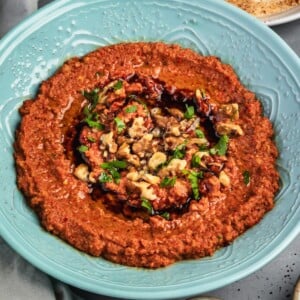 A bowl of muhammara dip garnished with walnuts, pomegranate molasses and parsley with a side of sliced cucumbers and pita.