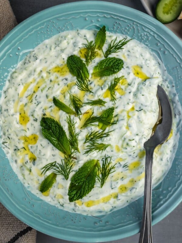 Tzatziki dip in a bowl garnished with mint, dill and a drizzle of olive oil with a side of pita and cucumber slices.