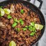 Pulled beef barbacoa with cilantro and lemon wedges as garnish in a braising dish.