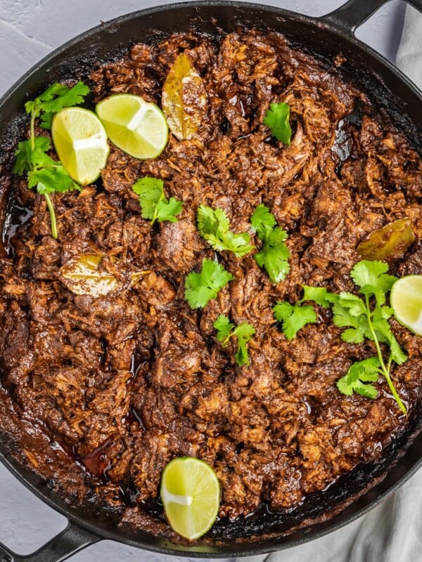 Pulled beef barbacoa with cilantro and lemon wedges as garnish in a braising dish.