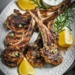 Grilled and marinated Greek lamb chops on a platter with rosemary and lemon wedges.