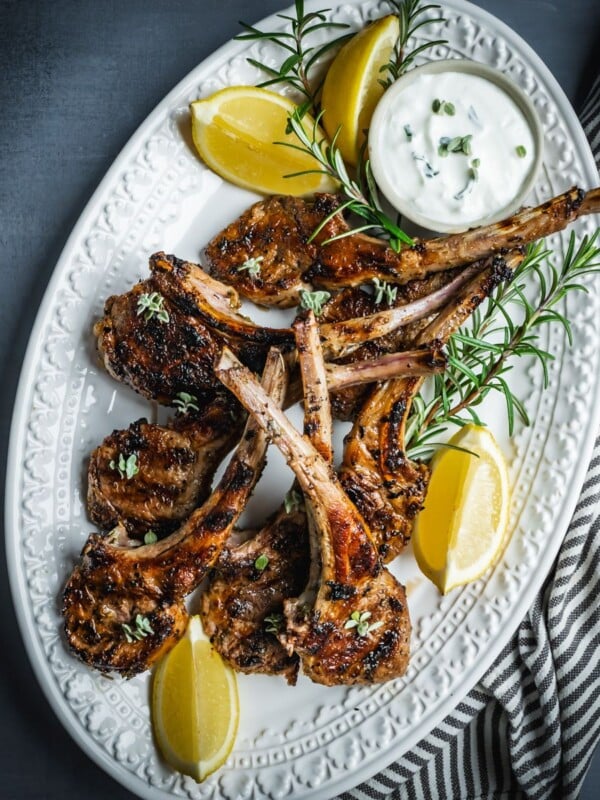 Grilled and marinated Greek lamb chops on a platter with rosemary and lemon wedges.