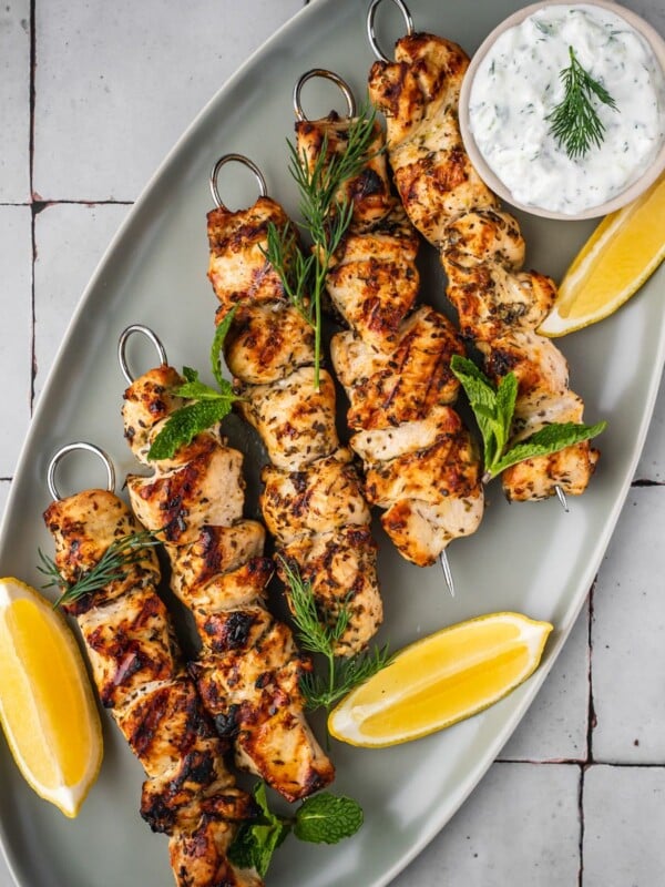 Chicken souvlaki skewers on a platter garnished with dill, mint, sliced lemons and a side of yogurt sauce.