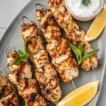 Greek chicken souvlaki skewers on a platter garnished with dill, mint, sliced lemons and a side of yogurt sauce.