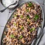 Platter with hashweh spiced rice with ground beef garnished with parsley and toasted nuts with a side of plain yogurt.