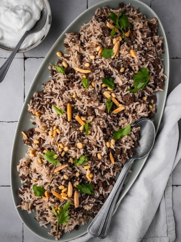 Platter with hashweh spiced rice with ground beef garnished with parsley and toasted nuts with a side of plain yogurt.