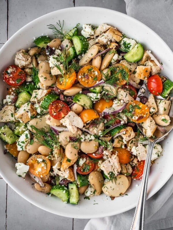 Greek chicken salad with butter beans, cherry tomatoes, feta and cucumbers garnished with fresh dill fronds in a large serving bowl.