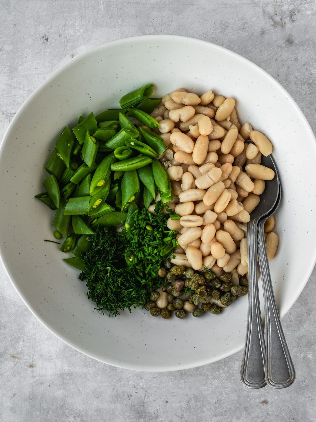 A bowl with canned white beans, chopped snap peas, capers and fresh herbs.