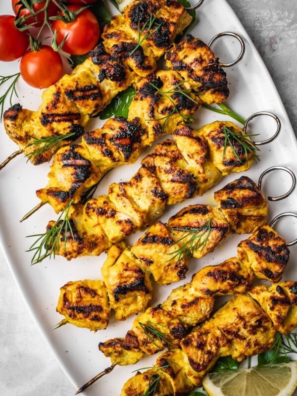 Grilled Joojeh Kabob chicken skewers with a golden char served on a white platter, garnished with fresh dill, cherry tomatoes, and lemon slices.