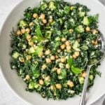 A bowl filled with a fresh salad made of kale, chickpeas, cucumber, herbs, and crumbled cheese. A spoon rests in the bowl.
