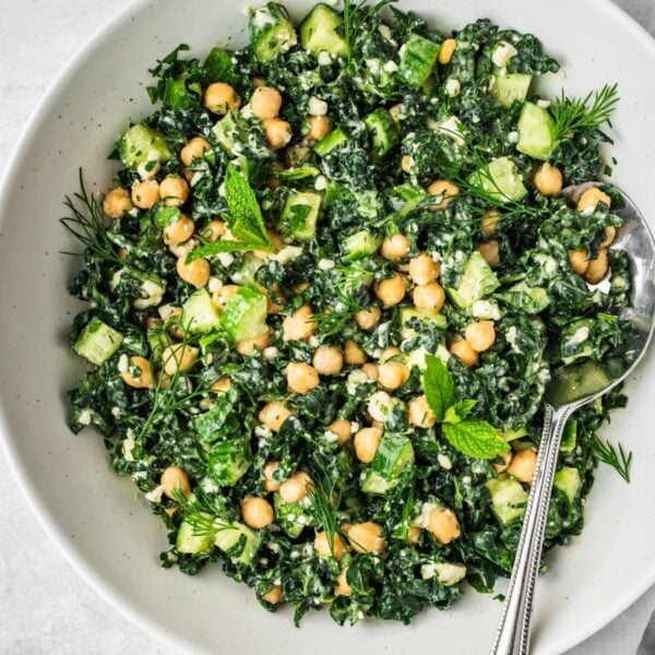 A bowl filled with a fresh salad made of kale, chickpeas, cucumber, herbs, and crumbled cheese. A spoon rests in the bowl.