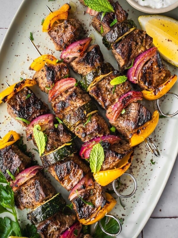 Middle Eastern Beef Shish Kebabs with yellow bell peppers, red onions, and zucchini on skewers, garnished with fresh mint and parsley. Served on a white platter with a lemon wedge and a small bowl of sauce.