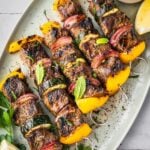 Middle Eastern Beef Shish Kebabs with yellow bell peppers, red onions, and zucchini on skewers, garnished with fresh mint and parsley. Served on a white platter with a lemon wedge and a small bowl of sauce.