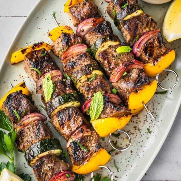 Middle Eastern Beef Shish Kebabs with yellow bell peppers, red onions, and zucchini on skewers, garnished with fresh mint and parsley. Served on a white platter with a lemon wedge and a small bowl of sauce.