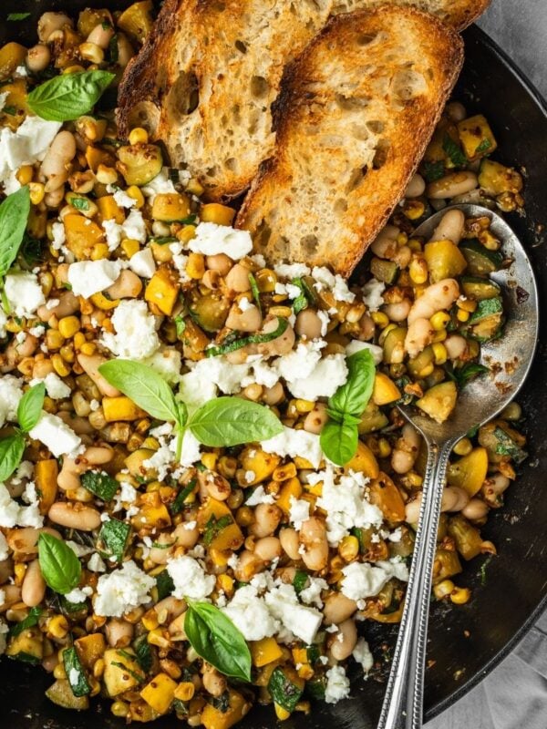 Sautee pan with corn, zucchini, white beans garnished with feta and basil leaves, served with a side of crust bread.