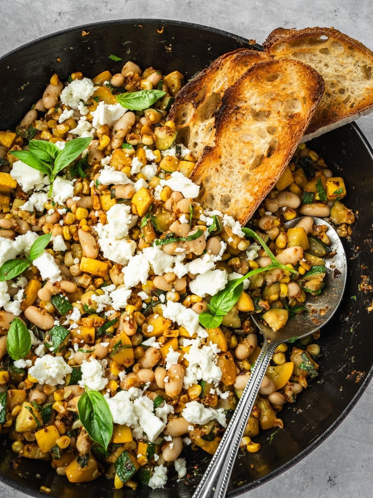 Saute pan with corn, zucchini, white beans garnished with feta and basil leaves, served with a side of crust bread.