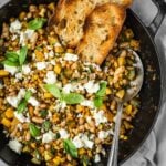 Sautee pan with corn, zucchini, white beans garnished with feta and basil leaves, served with a side of crust bread.