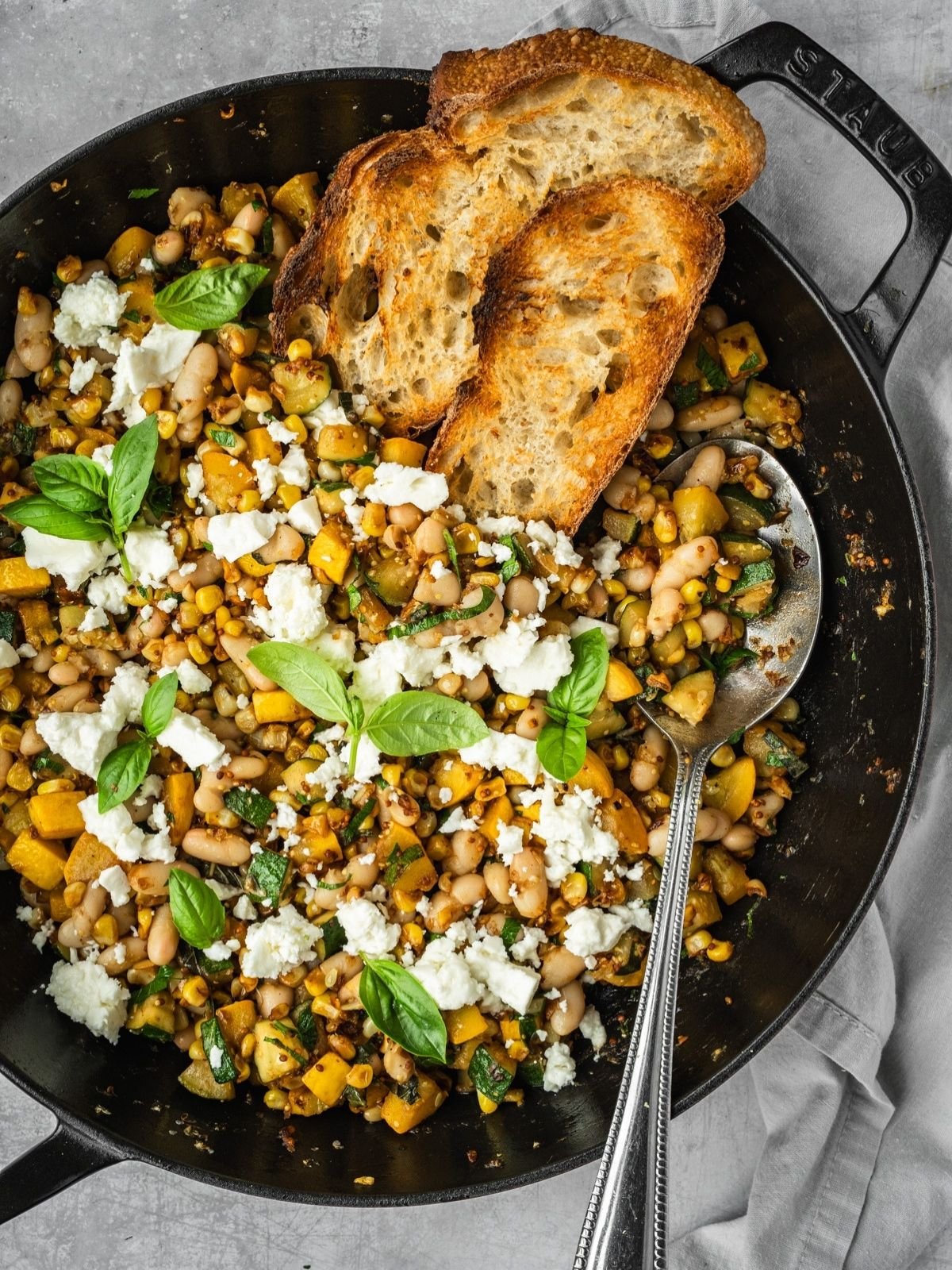 Sautee pan with corn, zucchini, white beans garnished with feta and basil leaves, served with a side of crust bread.
