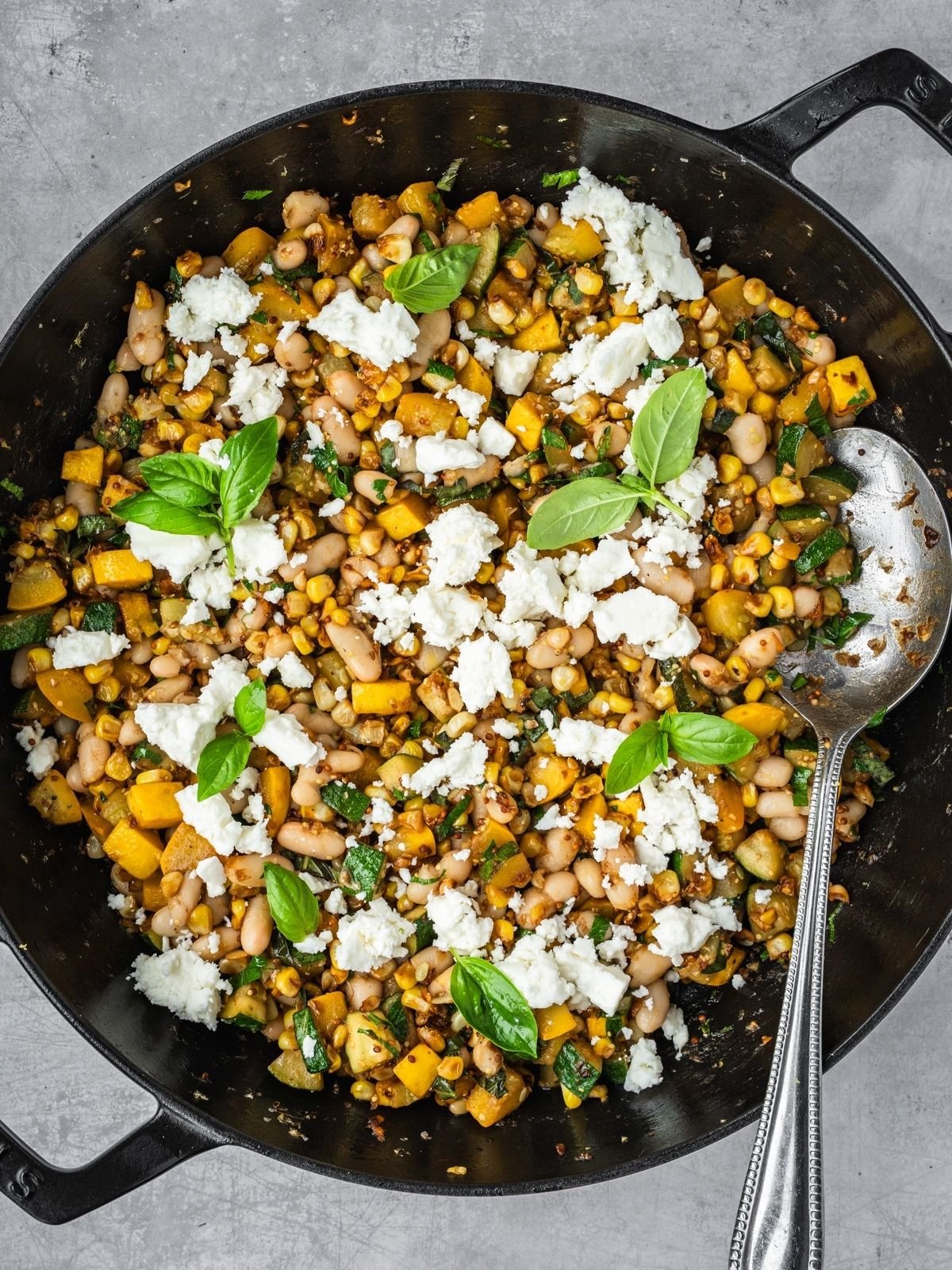 Sauté pan with corn, zucchini, white beans garnished with feta and basil leaves.