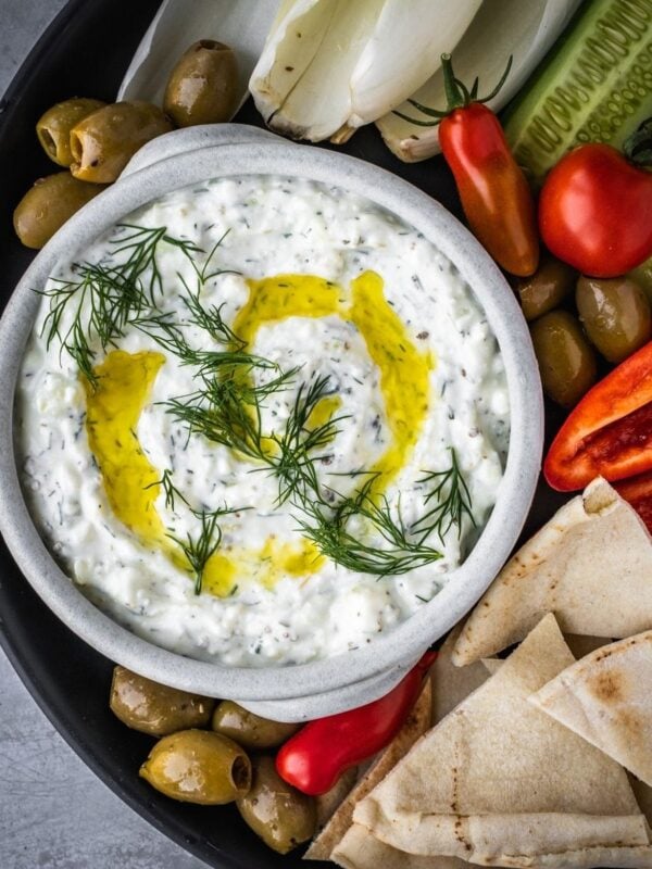 Feta yogurt dip garnished with fresh dill and olive oil on a platter with olives, tomatoes, sliced cucumbers and pita bread triangles.