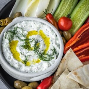 Feta yogurt dip garnished with fresh dill and olive oil on a platter with olives, tomatoes, sliced cucumbers and pita bread triangles.