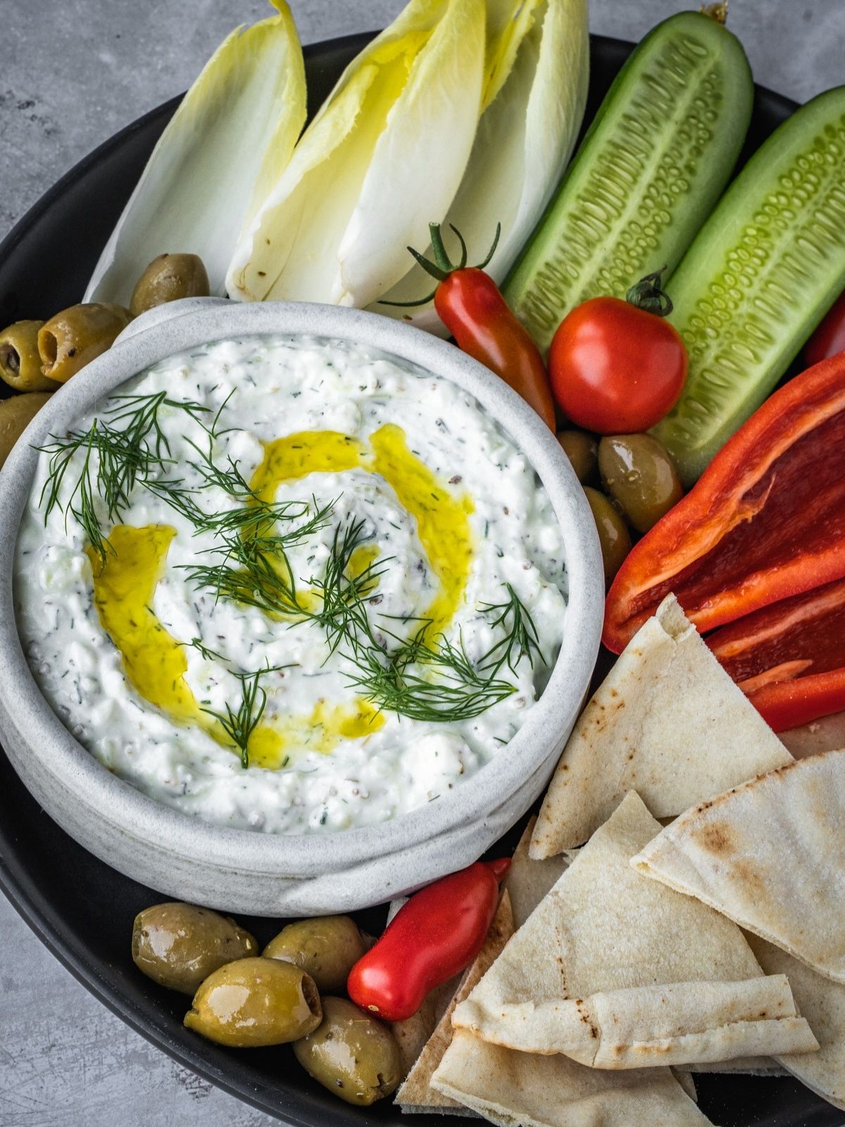 Feta yogurt dip garnished with fresh dill and olive oil on a platter with olives, tomatoes, sliced cucumbers and pita bread triangles.