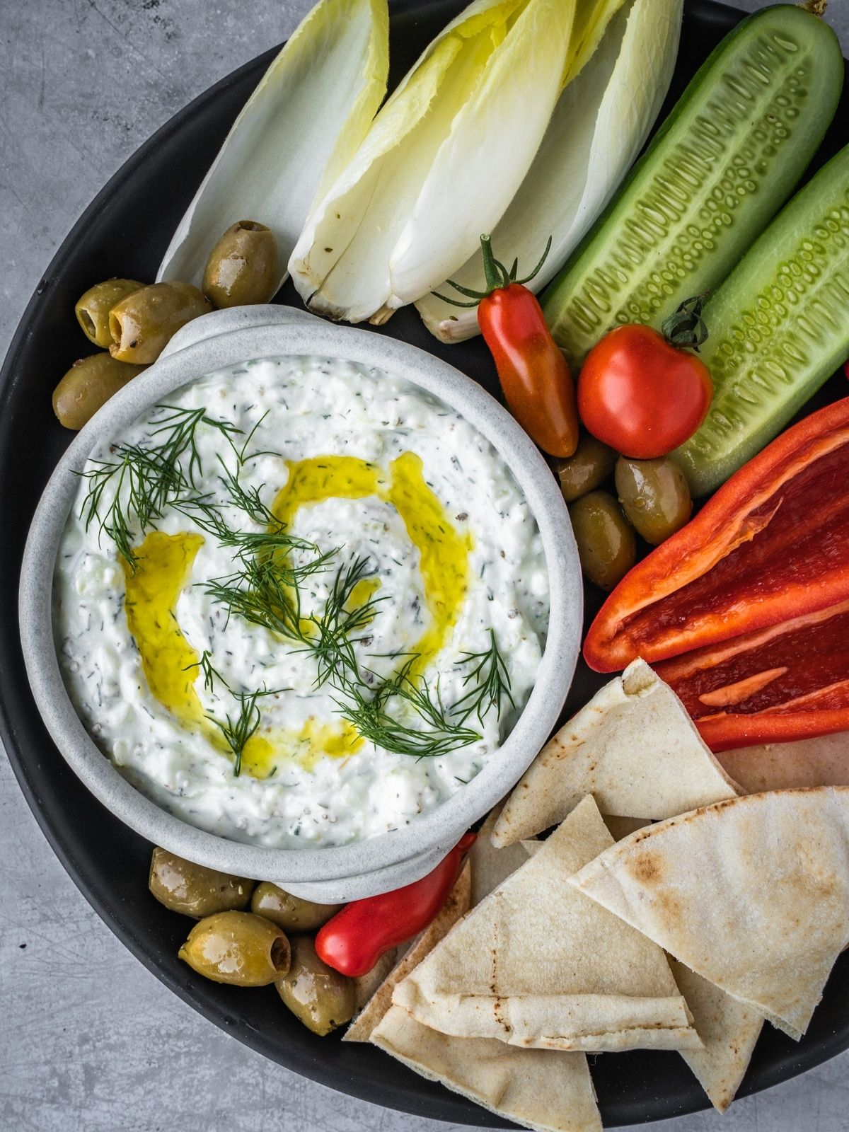 Feta yogurt dip garnished with fresh dill and olive oil on a platter with olives, tomatoes, sliced cucumbers and pita bread triangles.