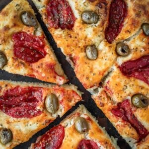 Focaccia Barese slices garnished with tomatoes, green olives and oregano on a wooden board.